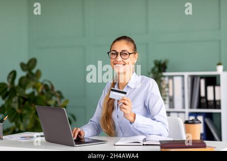 Porträt einer glücklichen Geschäftsfrau, die auf einem Laptop online einkaufen und eine Kreditkarte in der Hand hält, im Büroinnenraum lächelnd vor der Kamera lächelt und freien Platz hat Stockfoto