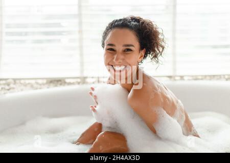 Porträt einer fröhlichen jungen Frau, die mit Schaum baden, die Kamera lächeln und sich morgens zu Hause in der heißen Badewanne entspannen. Schöne tausendjährige weibliche enjoyi Stockfoto