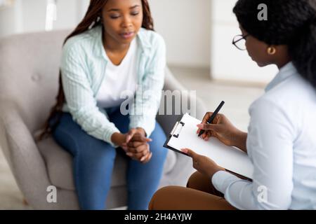 Gestresste schwarze weibliche Patientin, die Psychologin im medizinischen Büro konsultiert, selektiver Fokus. Psychiater hilft traurigen afroamerikanischen Frau mit nervös Stockfoto