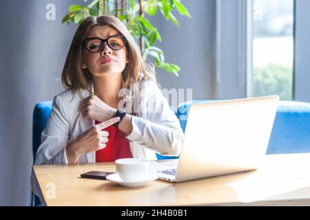 Porträt einer unglücklichen Frau in einer Brille, die auf die Smartwatch auf der Hand zeigt, düster verärgert aussieht, von Überstundenarbeit erschöpft ist und eine Pause braucht. Innenaufnahme, Café oder Bürohintergrund. Stockfoto