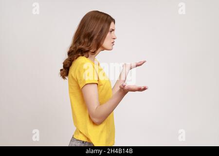 Was willst du von mir? Profilportrait von verwirrten jungen Weibchen mit braunem Haar in beiläufigen gelben T-Shirt mit sich ausbreitenden Händen, unbequeme Situation. Innenaufnahme des Studios isoliert auf grauem Hintergrund. Stockfoto