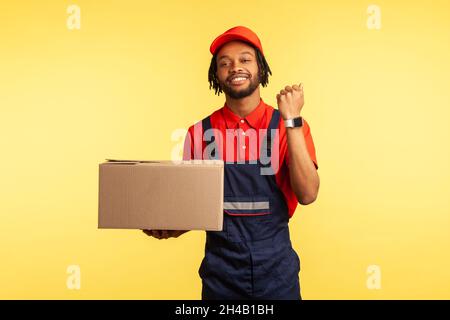 Angenehm aussehende Deliveryman in Overalls hält Papppaket und zeigt Armbanduhr, Lieferung in der Zeit, Versand und Frachttransport Service. Innenaufnahme des Studios isoliert auf gelbem Hintergrund. Stockfoto