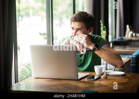 Porträt eines verärgerten Geschäftsmannes, der ein grünes T-Shirt trägt, vor dem Laptop sitzt, weint und seine Tränen mit Gewebe reinigt und Traurigkeit ausdrückt. Innenaufnahme in der Nähe des großen Fensters, Café-Hintergrund. Stockfoto