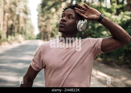 Afroamerikanischer Athlet, der Kopfhörer trägt, sich nach dem Laufen ausruhte, sich nach einem harten Training im Freien ausruhte, Fitness-Mann, der Musik mit Kopf zuhört Stockfoto