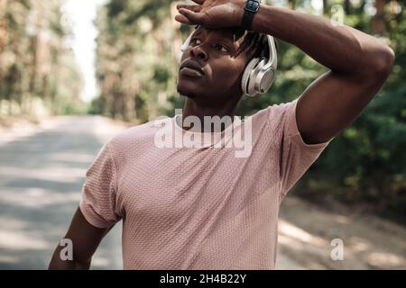 Afroamerikanischer Athlet, der Kopfhörer trägt, sich nach dem Laufen ausruhte, sich nach einem harten Training im Freien ausruhte, Fitness-Mann, der Musik mit Kopf zuhört Stockfoto