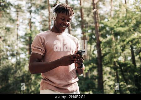 Fröhlicher, schwarzer Sportler, der Musik über Kopfhörer und Telefon hört, während er sich entspannt und im Park im Freien trainiert Stockfoto