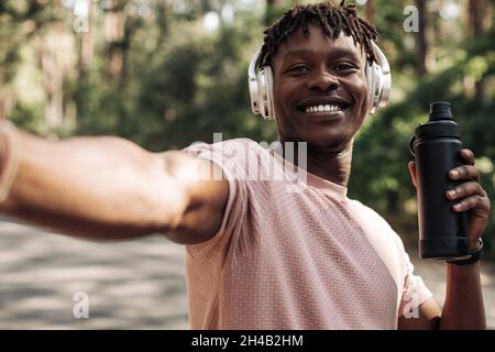 Porträt eines lächelnden afroamerikanischen Mannes mit Kopfhörern, der nach dem Joggen Selfie mit einer Wasserflasche im Park gemacht hat Stockfoto