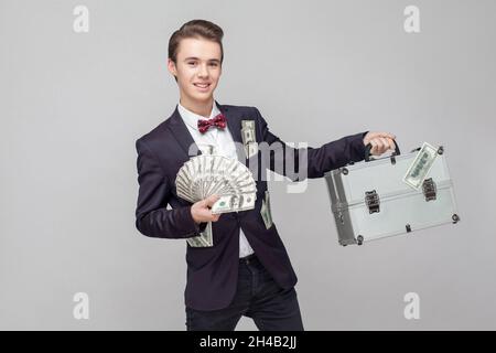 Porträt von fröhlich aufgeregt Millionär, Geschäftsmann mit stilvollen Frisur in schwarzem Anzug mit vielen Dollar in der Tasche zeigt Koffer und Haufen Geld. Indoor-Studio auf grauem Hintergrund isoliert Stockfoto