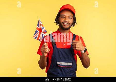 Porträt eines positiven Arbeiters in blauen Overalls, der die britische Flagge hält und den Daumen nach oben zeigt, der die Kamera zufrieden anschaut. Innenaufnahme des Studios isoliert auf gelbem Hintergrund. Stockfoto