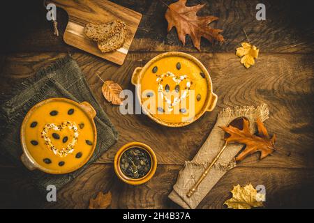 Zu Schüsseln mit Kürbissuppe mit Creme und Samen auf rustikalem Metall- und Holzhintergrund, verziert mit Herbstblättern, Draufsicht Stockfoto