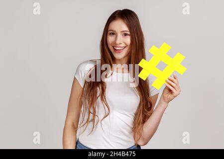 Lächelnd schöne Frau mit gelbem Hashtag-Symbol, macht wichtiges Thema populär, setzt Trends im Internet, trägt weißes T-Shirt. Innenaufnahme des Studios isoliert auf grauem Hintergrund. Stockfoto