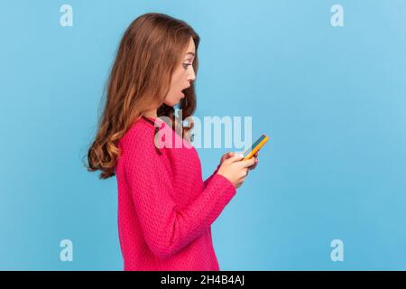 Seitenansicht einer staunenden Frau, die einen rosa Pullover trägt, schockiert auf das Mobiltelefon schaut, aktuelle Nachrichten in sozialen Netzwerken liest, Anwendung. Innenaufnahme des Studios isoliert auf blauem Hintergrund. Stockfoto