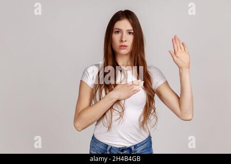 Porträt einer sehr verantwortungsbewussten und ehrlichen Frau, die Versprechen gibt, feierliches Gelübde in zeremonieller Tradition mit erhobener Hand und weißem T-Shirt. Innenaufnahme des Studios isoliert auf grauem Hintergrund. Stockfoto