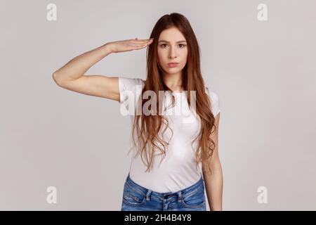 Porträt einer verantwortungsbewussten, seriöse Frau mit dunklen Haaren, die den Kommandanten grüßen, mit gehorsamem Ausdruck und weißem T-Shirt. Innenaufnahme des Studios isoliert auf grauem Hintergrund. Stockfoto