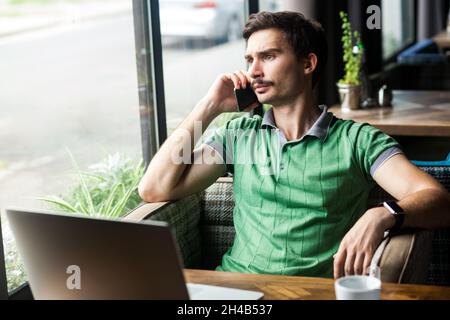 Porträt eines freiberuflichen Mannes, der grünes T-Shirt trägt, Mobiltelefon spricht, Probleme diskutiert, wegschaut, mit einem Geschäftspartner spricht. Innenaufnahme in der Nähe des großen Fensters, Café-Hintergrund. Stockfoto