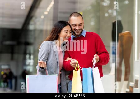 Verkaufssaison, riesige Rabatte. Glückliches junges multirassisches Paar, das in farbenfrohe Einkaufstaschen schaut, zufrieden mit ihren Einkäufen, zusammen stehend o Stockfoto