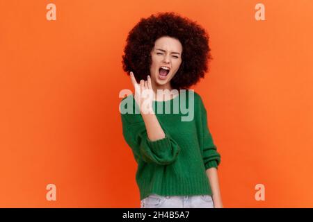 Aufgeregt enthusiastische Frau mit Afro-Frisur trägt grünen lässigen Stil Pullover mit Rock and Roll Geste. Schreiend begeistert, Indoor Studio Schuss isoliert auf orangefarbenem Hintergrund. Stockfoto