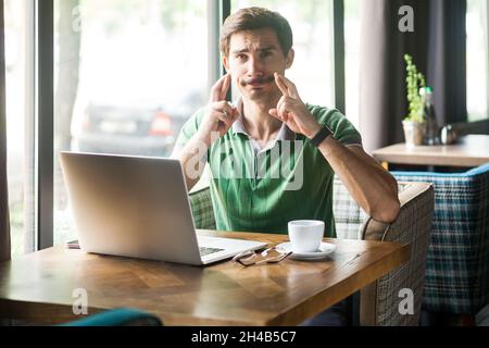 Porträt eines Mitarbeiters mit grünem T-Shirt, gekreuzten Fingern und dem Wunsch nach Glück, in Erwartung des Erfolgs, Online-Arbeit am Laptop, Innenaufnahme in der Nähe eines großen Fensters, Café-Hintergrund. Stockfoto