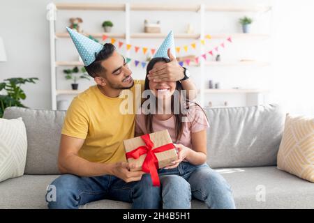 Hübscher junger arabischer Kerl, der seine Freundinnen mit den Augen bedeckt, eine Geburtstagsüberraschung macht und ihr zu Hause ein verpacktes Geschenk gibt. Millennial verschiedene Paar feiern Stockfoto