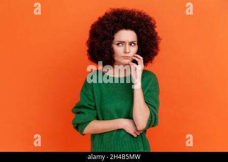 Nachdenkliche Frau mit Afro-Frisur trägt grünen lässigen Stil Pullover denken, wegschauen, verwirrt verwirrt Gesichtsausdruck. Innenaufnahme des Studios isoliert auf orangefarbenem Hintergrund. Stockfoto