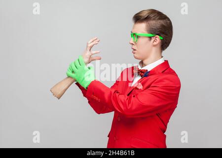 Portrait von bizarren lustigen Gentleman mit Brillen in rotem Smoking und grünen Handschuhen, die erschrocken aussehen und Zombie Hand halten, um ihn zu fangen. studio Shot isoliert auf grauem Hintergrund Stockfoto