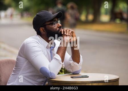 Porträt eines jungen Mannes, der an einem Tisch in einem Straßencafé sitzt. Er trägt ein weißes Hemd und eine Mütze Stockfoto