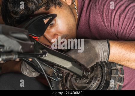 Nahaufnahme eines jungen lateinischen Mechanikers, der die Verkabelung, die zum Rad eines Elektrorollers in seiner Werkstatt geht, sorgfältig überprüft Stockfoto