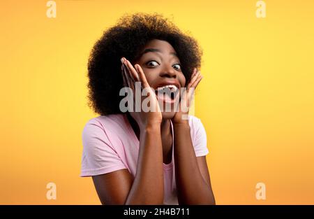 Wow-Angebot. Überglücklich afroamerikanische Dame, die begeistert auf die Kamera schaut und das Gesicht auf gelbem Hintergrund berührt, Studioaufnahme. OMG Emotion Konzept Stockfoto