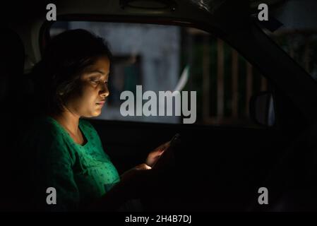 Indische Frau, die ihr Handy in einem Auto in der Dunkelheit sitzend ansieht Stockfoto