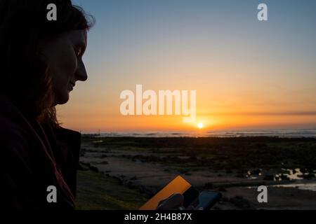 Frau, die Mobiltelefon benutzt, während die Sonne am West Runton Beach untergeht Stockfoto