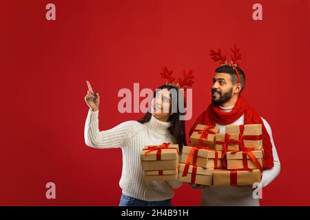 Urlaubsangebot und Promo. Glückliches junges arabisches Paar, das auf freien Platz für Ihre Werbung auf rotem Hintergrund zeigt und schaut, Mann und Frau tragen Stockfoto