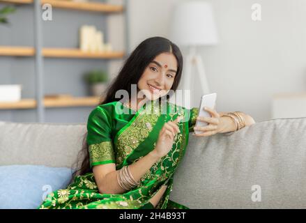 Porträt einer schönen indischen Frau im Sari-Kleid mit Mobiltelefon auf dem Sofa zu Hause. Charmante junge Dame aus dem Osten in ethnischen Kleidern sucht im Internet, chec Stockfoto
