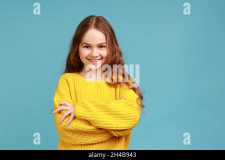 Porträt von niedlichen kleinen Mädchen stehen mit gefalteten Armen und Blick auf die Kamera mit glücklichem Ausdruck, tragen gelben lässigen Stil Pullover. Innenaufnahme des Studios isoliert auf blauem Hintergrund. Stockfoto