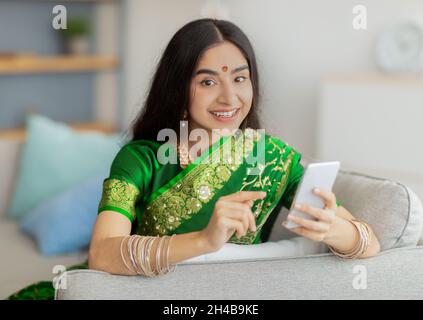 Charmante indische Dame im traditionellen Saree sitzt auf dem Sofa mit Handy, macht einen Videoanruf an einen Freund von zu Hause. Millennial Eastern Frau in ethnischen cl Stockfoto