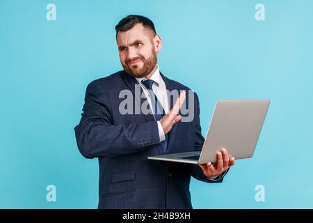 Portrait eines attraktiven bärtigen jungen Mannes, der einen Anzug im offiziellen Stil trägt, der die Hand dehnt, nicht sehen will, weg schaut, ekelhafte Inhalte, Innenaufnahme des Studios auf blauem Hintergrund isoliert. Stockfoto