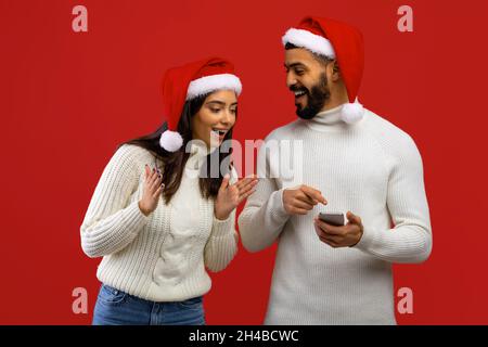 Tolle Weihnachtsnachrichten. Überglücklich arabische Ehegatten in Santa-Hüten, empfangen Nachricht, Mann zeigt Smartphone zu Frau, posiert zusammen über roten Studio-Backgro Stockfoto