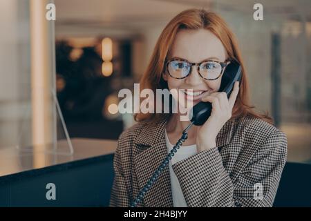 Büroangestellte in einer Brille, die mit ihrer Kollegin über das Festnetztelefon spricht und die Kamera anlächelt Stockfoto