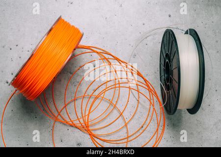 Kunststofffilamente für 3D-Drucker. Spulen aus orangefarbenen und weißen thermoplastischen Drähten für 3D-Druck aus nächster Nähe auf grauem Steingrund, flach liegend Stockfoto