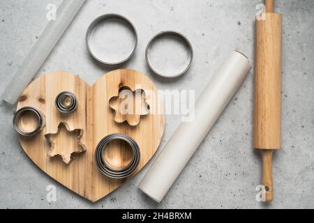 Ausstecher für Plätzchenteig, Pergamentpapier Nahaufnahme auf herzförmigem Holzbrett auf grauem Steingrund, flach ausgelegt Stockfoto