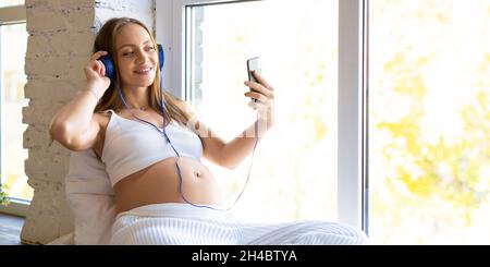 Eine Schwangere mit blauen Kopfhörern sitzt zu Hause auf dem Fensterbrett. Sie hat Online-Videoanruf – Online-Konsultation mit einem Arzt oder Online-Meeting wi Stockfoto