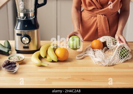 Hände einer jungen Hausfrau, die frisches Obst aus dem Beutel nimmt, während sie am Tisch steht und Zutaten für Smoothie zubereitet Stockfoto