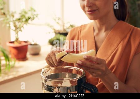 Junge Frau hackt geschälte Banane über den elektrischen Mixer, während sie hausgemachten Smoothie für das Familienfrühstück zubereitet Stockfoto