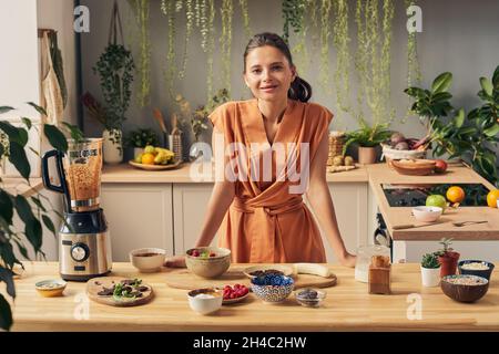 Glückliche junge Hausfrau, die Sie anschaut, während sie am Küchentisch mit Smoothie-Zutaten in Schüsseln und elektrischem Mixer steht Stockfoto