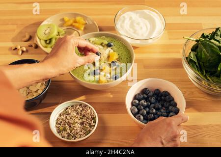 Junge Frau, die Smoothie mit grünem Gemüse in einer Schüssel mit frischen Heidelbeeren schmückt, bevor sie es zum Frühstück servierte Stockfoto