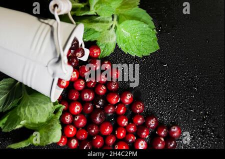 Preiselbeeren mit Minze werden aus dem weißen Eimer geströmt. Dunkler Hintergrund. Speicherplatz kopieren. Alles ist mit Wassertröpfchen bedeckt. Stockfoto