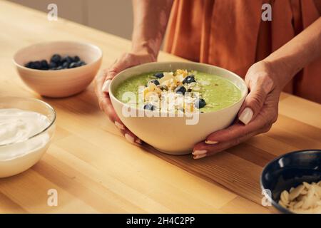 Hände einer Frau halten Schüssel mit frischen hausgemachten Smoothie mit Heidelbeeren, Müsli, Orangen und Mandelflocken dekoriert Stockfoto