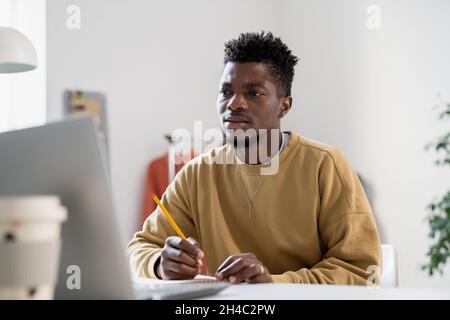 Angespannter junger Mann mit Bleistift, der während des Online-Unterrichts Notizen macht und dem Lehrer zuhört, auf den Laptop-Bildschirm schaut Stockfoto