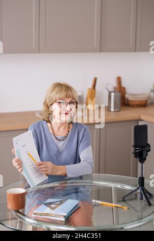 Glückliche blonde reife Lehrerin zeigt auf Dokument, während sie dem Online-Publikum während des Online-Trainings Informationen erklärt Stockfoto