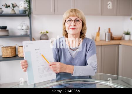 Selbstbewusste blonde Frau zeigt auf Finanzpapier und erklärt das Thema am Küchentisch vor der Kamera Stockfoto