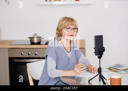 Selbstbewusste, reife Trainerin, die während des Online-Trainings in der Smartphone-Kamera sitzt, während sie am Küchentisch sitzt Stockfoto